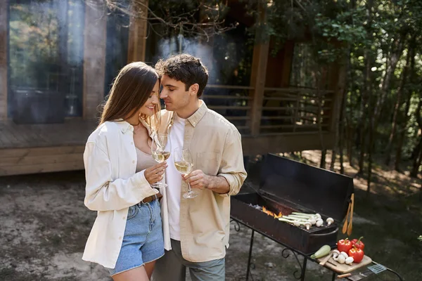Позитивная пара держит вино возле размытого барбекю и дома отдыха на заднем плане на открытом воздухе — стоковое фото