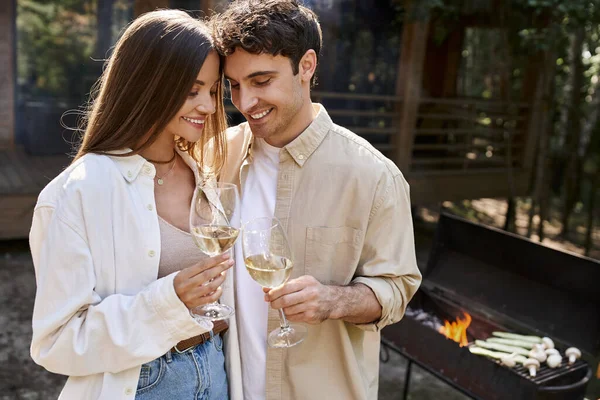 Sonriente pareja tostadas con vino cerca de la parrilla borrosa y casa de vacaciones en el fondo al aire libre — Stock Photo