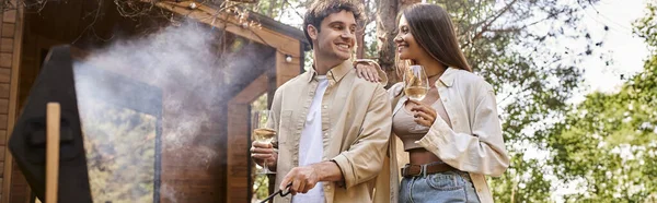Donna sorridente che tiene il vino vicino al fidanzato, alla griglia e alla casa di vacanza sullo sfondo, banner — Foto stock