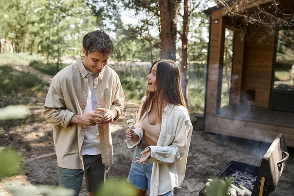 Lächelnde Frau hält Wein in der Hand und spricht mit Freund in der Nähe von Grill und Ferienhaus im Hintergrund — Stockfoto