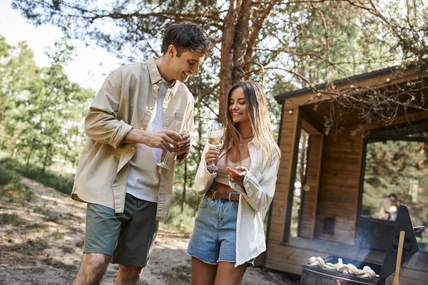 Couple souriant en vêtements décontractés parlant et tenant du vin près du gril et de la maison de vacances en arrière-plan — Photo de stock