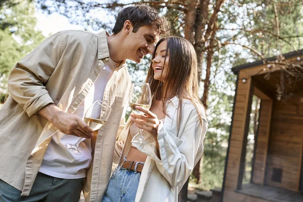 Pareja positiva en ropa casual sosteniendo vino durante el picnic cerca de la casa de vacaciones en el fondo - foto de stock