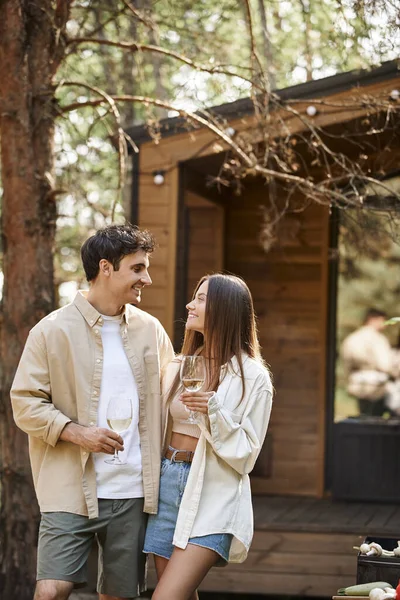 Positive brunette couple holding wine during picnic near barbecue and vacation house at background — Stock Photo