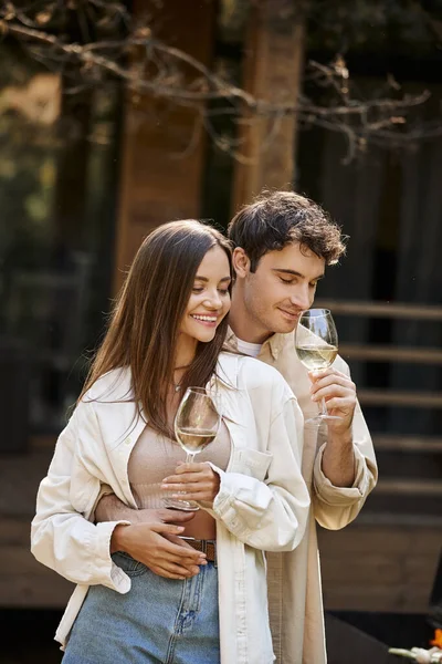 Lächelnder Mann hält Wein in der Hand und umarmt Freundin neben Grill und verschwommenem Ferienhaus im Hintergrund — Stockfoto