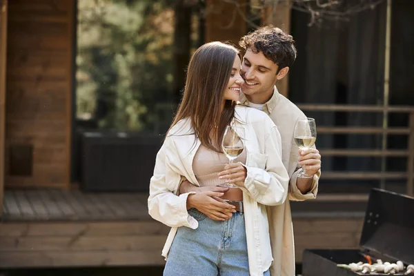 Sorrindo homem abraçando namorada e segurando vinho perto de churrasco e casa de férias borrada no fundo — Fotografia de Stock
