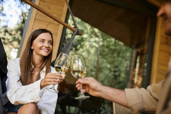 Donna sorridente brindare vino con fidanzato offuscato e seduto in amaca vicino casa vacanza — Foto stock