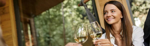 Felice bruna donna tostare vino con fidanzato mentre seduto in amaca vicino alla casa estiva, striscione — Foto stock