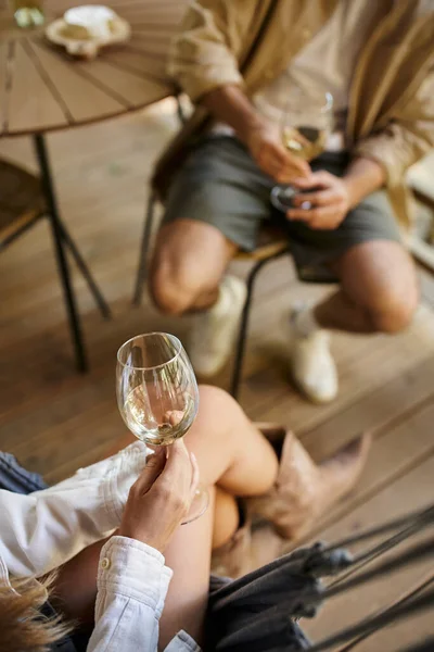 Vista recortada de la mujer joven sosteniendo el vino y sentado en la hamaca cerca de novio y casa de verano - foto de stock