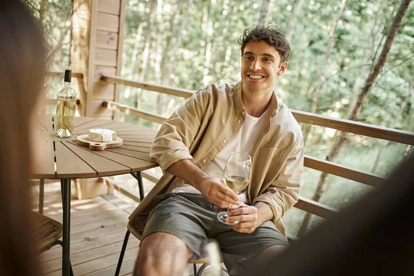 Smiling brunette man holding wine near cheese and blurred girlfriend on terrace of vacation house — Stock Photo