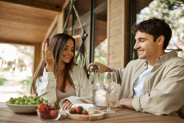 Smiling man pouring wine near girlfriend with strawberry on terrace of vacation house at background — Stock Photo