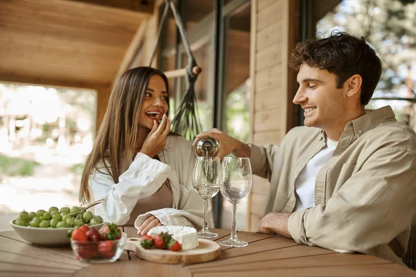 Lächelnder Mann schenkt Wein ein, während Freundin Erdbeere auf der Terrasse ihres Ferienhauses isst — Stockfoto
