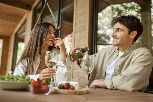 Mulher sorrindo comendo morango perto de namorado com vinho no terraço da casa de férias de madeira — Fotografia de Stock