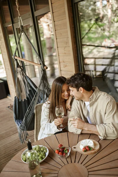 Positive woman hugging boyfriend near wine and food on terrace of wooden summer house — Stock Photo