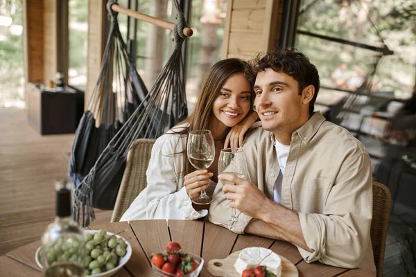 Feliz pareja romántica sosteniendo vino cerca de frutas y queso en la terraza de la casa de vacaciones durante el verano - foto de stock