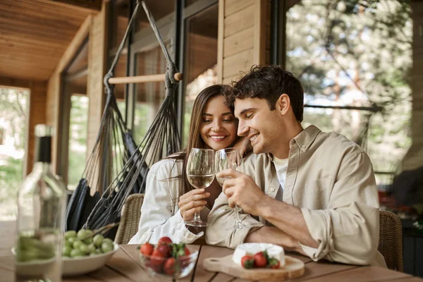 Sonriente pareja romántica tintineo con vino cerca de la comida borrosa en la terraza de la casa de vacaciones de madera - foto de stock