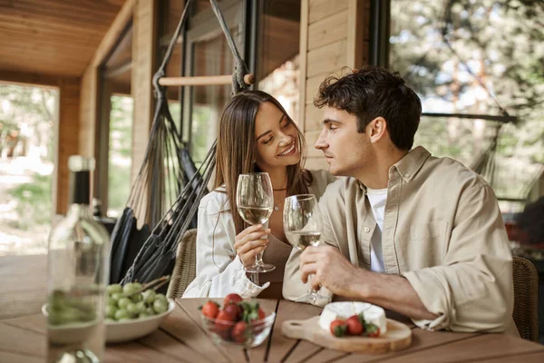 Mujer sonriente sosteniendo el vino y mirando al novio cerca de la comida borrosa en la terraza de la casa de verano - foto de stock