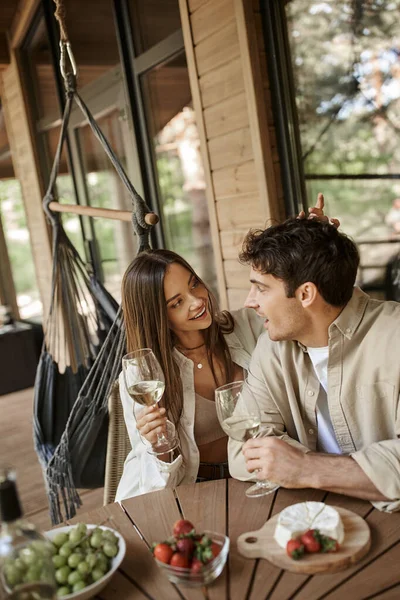 Mujer sonriente con vino hablando y tocando novio cerca de la comida en la terraza de la casa de vacaciones - foto de stock
