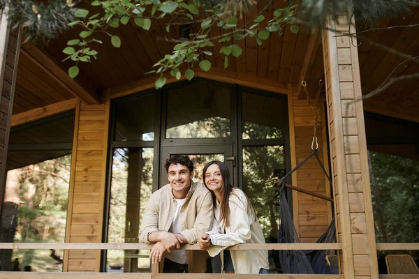 Улыбающаяся романтическая пара смотрит в камеру, стоя на террасе деревянного домика на открытом воздухе — стоковое фото