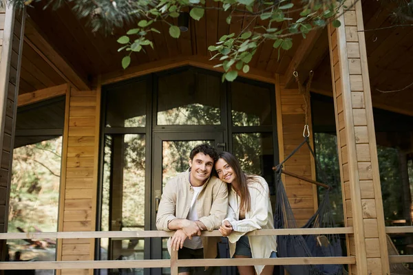 Cheerful brunette couple looking at camera while standing on terrace of wooden summer house outdoors — Stock Photo