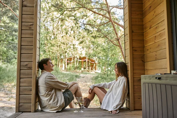 Vue latérale du couple romantique positif assis près du vin sur le porche de la maison d'été en bois à l'extérieur — Photo de stock
