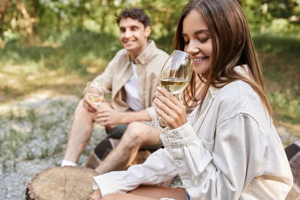 Sonriente joven mujer sosteniendo un vaso de vino y sentado cerca borrosa novio al aire libre - foto de stock