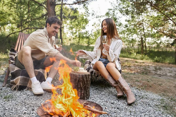 Cheerful couple with wine talking while sitting on deck chairs near food and firewood outdoors — Stock Photo