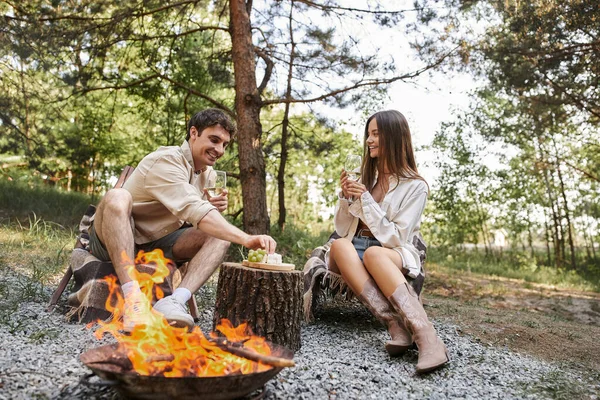 Positives Paar mit Wein und Essen sitzt auf Liegestühlen in der Nähe von Brennholz beim Picknick im Freien — Stockfoto