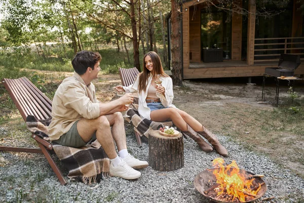 Une jeune femme positive tenant du vin et parlant à son petit ami près de la nourriture et du bois de chauffage à l'extérieur — Photo de stock