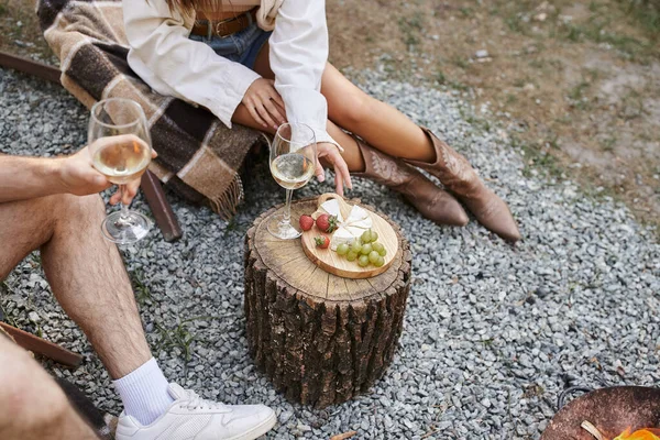 Vista cortada de casal romântico segurando vinho perto de frutas, queijo e lenha ao ar livre durante o verão — Fotografia de Stock