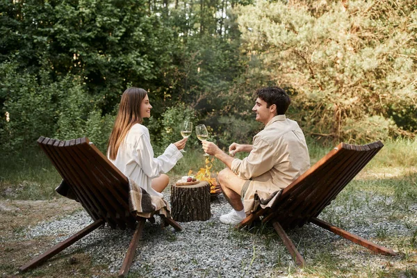 Vista lateral do casal romântico sorridente segurando vinho perto de comida e lenha durante o piquenique ao ar livre — Fotografia de Stock