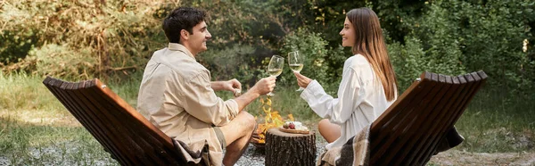 Side view of smiling couple holding wine near food and firewood during vacation, banner — Stock Photo