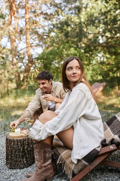 Mujer morena sentada en la silla de cubierta cerca de novio con vino y comida durante el picnic al aire libre - foto de stock