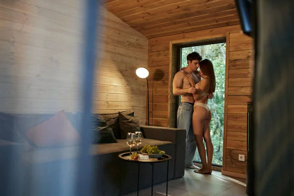 Mulher sensual em roupa interior abraçando namorado sem camisa perto de comida e vinho em casa de férias — Fotografia de Stock