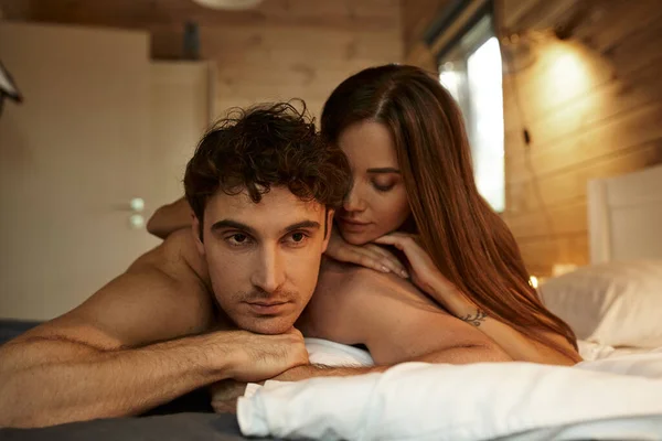 Чувственная татуированная женщина, лежащая на парне без рубашки на кровати в домике для отдыха вечером — стоковое фото