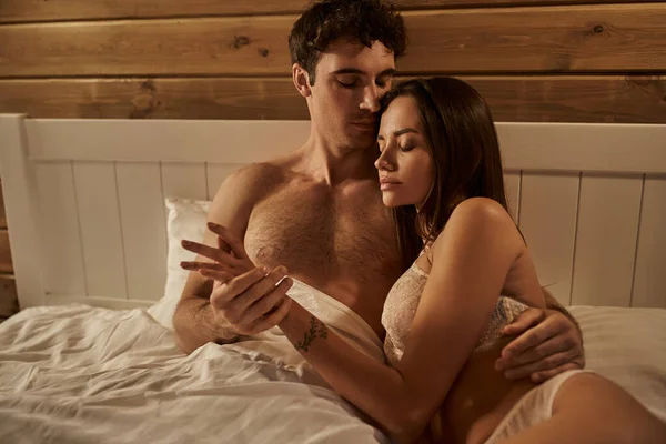 Homme sexy toucher la main de petite amie tatouée en lingerie sur le lit dans la maison de vacances — Photo de stock