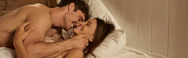Sonriente y sexy hombre tocando atractiva novia mientras está acostado en la cama en la casa de vacaciones, pancarta - foto de stock