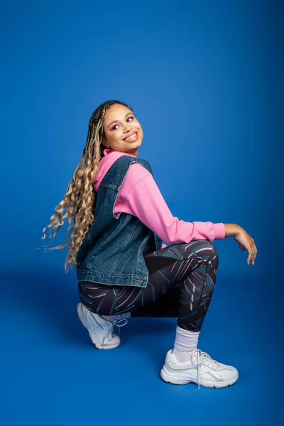 Happy african american woman with dreadlocks sitting and posing on blue background, sporty outfit — Stock Photo