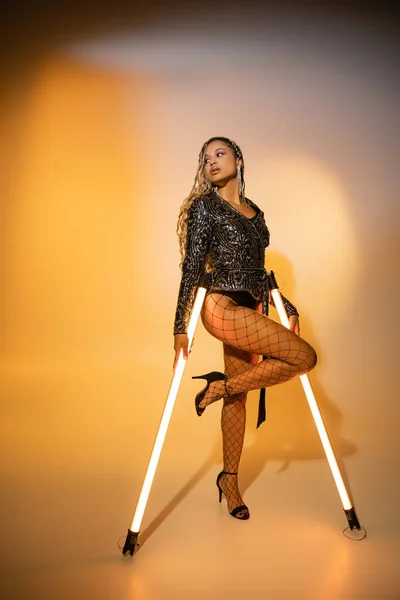 Bold style, sexy african american woman in fishnet tights posing with fluorescent lamps on yellow — Stock Photo