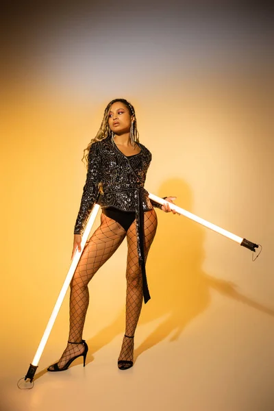 Style sexy, femme afro-américaine en collants résille et talons hauts posant avec des lampes fluorescentes — Photo de stock