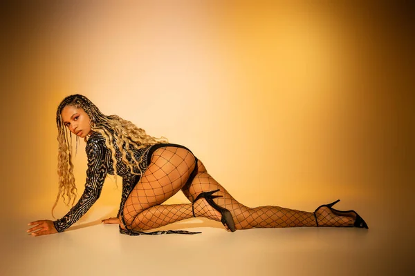Sexy african american model with tattoo crawling on yellow background, fishnet tights and high heels — Stock Photo