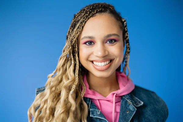 Alegre mujer afroamericana en sudadera con capucha rosa y chaleco vaquero sobre fondo azul, modelo de piel oscura — Stock Photo
