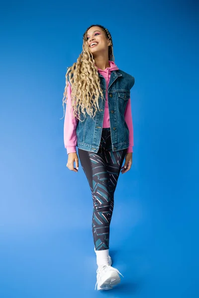 Toute la longueur de femme sportive afro-américaine souriant, regardant loin sur le bleu, gilet en jean et leggings — Photo de stock