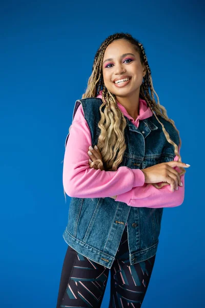 Joyful african american woman in pink hoodie and denim vest on blue background, bold makeup — Stock Photo
