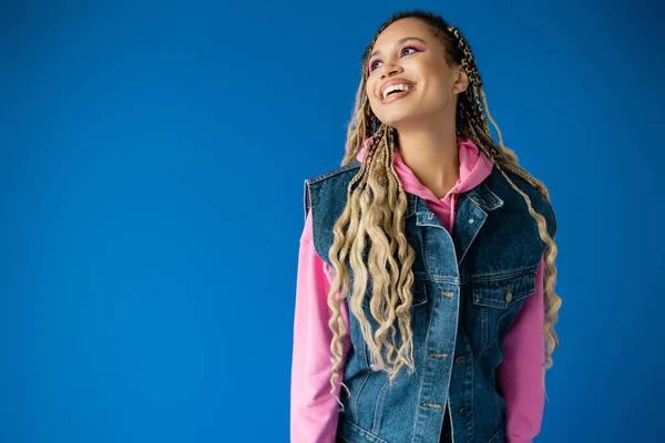 Щаслива афроамериканська жінка в рожевому светрі і джинсовому жилеті, посміхаючись на синьому фоні, сміливий макіяж — стокове фото