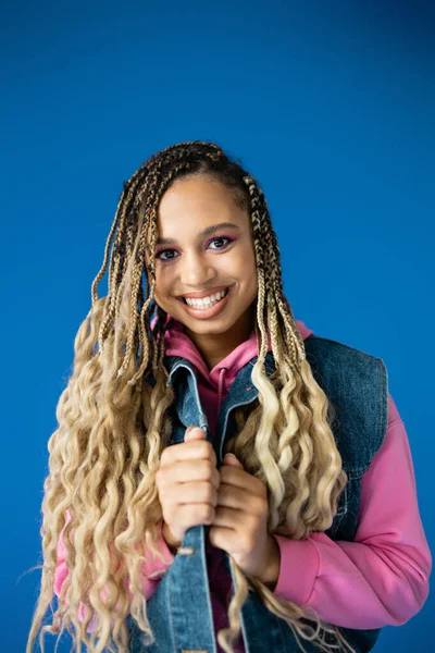 Happy african american woman in pink hoodie and denim vest smiling on blue background, dreadlocks — Stock Photo