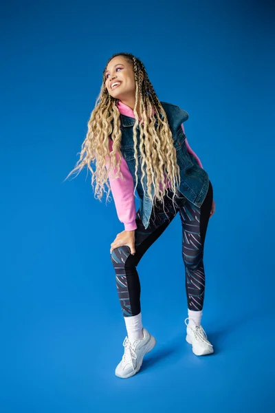 Full length, happy african american woman in pink hoodie and denim vest posing on blue background — Stock Photo