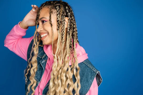 Positive african american woman in pink hoodie and denim vest smiling on blue background, joy — Stock Photo