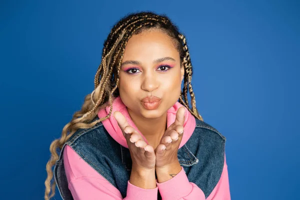 Attractive african american woman sending air kiss on blue background, denim vest and pink hoodie — Stock Photo