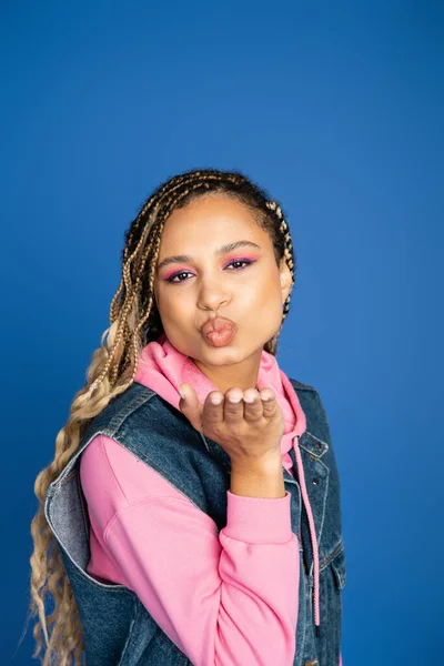Beautiful african american woman sending air kiss on blue background, denim vest and pink hoodie — Stock Photo