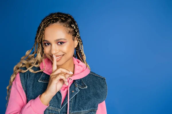 Happy african american woman with dreadlocks showing hush sign, blue background, finger near lips — Stock Photo